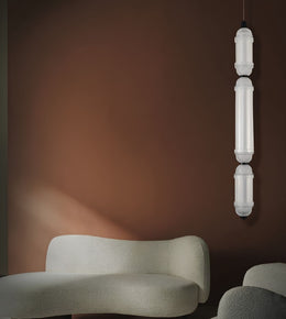 A modern vertical pendant light with three stacked cylindrical frosted white glass shades hangs against a warm brown wall above a curved cream-colored upholstered bench.