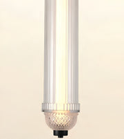 A modern cylindrical pendant light fixture with a ribbed clear acrylic shade, a glowing warm white vertical tube, a textured clear glass bowl, and a black top cap, shown against a plain cream background.