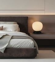 A modern bedroom scene featuring a bed with white linens and a dark brown upholstered headboard and footboard, alongside a floating nightstand holding a glowing spherical table lamp.