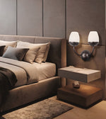 A modern bedroom scene featuring a bed with neutral-toned bedding, a matching upholstered headboard, a floating nightstand, and a dual-arm wall sconce with frosted white shades mounted on a textured taupe wall.