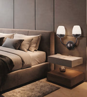 A modern bedroom scene featuring a bed with neutral-toned bedding, a matching upholstered headboard, a floating nightstand, and a dual-arm wall sconce with frosted white shades mounted on a textured taupe wall.