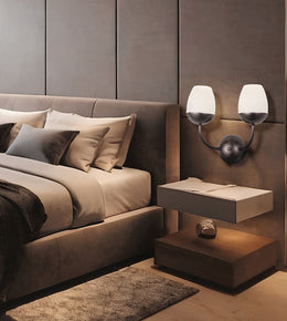 A modern bedroom scene featuring a bed with neutral-toned bedding, a matching upholstered headboard, a floating nightstand, and a dual-arm wall sconce with frosted white shades mounted on a textured taupe wall.
