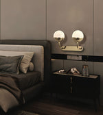 A modern wall-mounted double light fixture with spherical frosted white shades and brushed gold arms, installed above a dark nightstand in a contemporary bedroom setting.