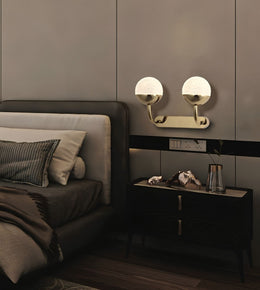 A modern wall-mounted double light fixture with spherical frosted white shades and brushed gold arms, installed above a dark nightstand in a contemporary bedroom setting.