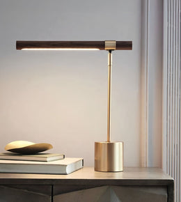 A modern table lamp with a horizontal dark wood head, a slim gold metal stem, and a cylindrical gold base, shown lit on a tabletop next to stacked books.