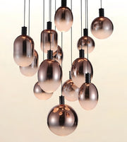 A cluster of modern pendant lights with metallic rose gold glass globes and teardrop shapes hanging from black cords against a warm off-white background.