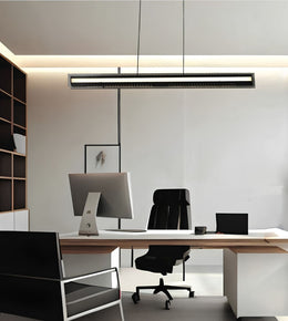 A modern minimalist office setup featuring a long, slim rectangular LED pendant light hanging over a light wood desk with a white computer monitor, black office chair, and laptop.