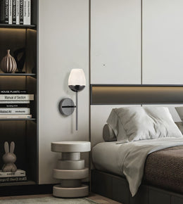 A modern wall-mounted sconce with a white frosted glass shade and a dark metal base, mounted on a bedroom wall next to a bed and a small round side table.