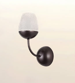 A modern wall-mounted sconce with a dark bronze-finished metal body, featuring a curved arm and a textured frosted glass shade, shown against a light beige background.