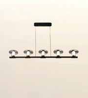 A modern 5-light linear ceiling chandelier with a matte black horizontal bar, clear glass dome shades, and exposed white bulb sockets, suspended by thin cables from a rectangular ceiling canopy.