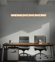 A modern office scene with a long horizontal linear pendant light glowing above a wooden desk, two office chairs, and a gray wall with strong sunlight shadows.
