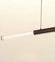 A modern linear pendant light with a ribbed clear glass shade and a dark brown metal body, suspended by a thin black cord against a plain off-white background.
