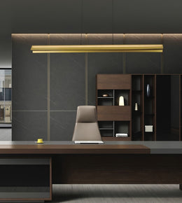 Modern office interior featuring a dark wood desk, a beige upholstered chair, a black shelving unit, and a long horizontal gold pendant light against a dark gray paneled wall.