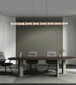 A modern linear pendant light with a textured, segmented wood-look finish hangs over a long conference table with chairs in a minimalist office setting.