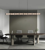 A modern linear pendant light with a textured, segmented wood-look finish hangs over a long conference table with chairs in a minimalist office setting.