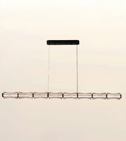 A modern linear chandelier with a textured metallic silver horizontal bar suspended by two thin cables from a black rectangular ceiling mount, shown against a plain light beige background.