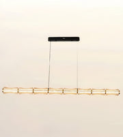 A modern linear pendant light fixture designed to resemble a segmented bamboo pole, featuring a warm gold-toned metallic finish and a sleek black ceiling canopy, suspended against a plain off-white background.