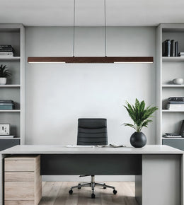 A modern minimalist home office setup featuring a long rectangular wooden pendant light hanging above a white desk with a black office chair. The room is styled symmetrically with light gray shelving units on either side containing books and plants.