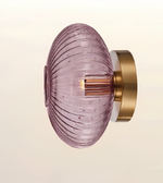A modern wall-mounted light fixture featuring a ribbed, translucent dusty purple glass shade and a brushed gold metal wall mount, shown against a clean off-white background.