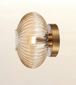 A modern wall-mounted light fixture with a ribbed, ribbed glass shade that glows with a warm light, attached to a brushed brass circular wall plate.