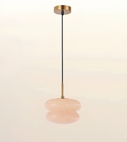 A modern pendant light with a brushed brass ceiling canopy and downrod, a black cord, and a double-tiered, rounded blush pink glass shade, shown against a warm off-white background.