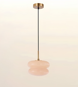 A modern pendant light with a brushed brass ceiling canopy and downrod, a black cord, and a double-tiered, rounded blush pink glass shade, shown against a warm off-white background.