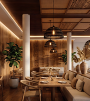 A modern restaurant interior with wooden ceiling panels, hanging black pendant lights, a wooden dining table, beige upholstered seating, and large potted plants.