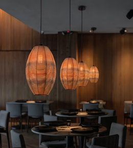 Three hanging pendant lights with woven wicker baskets in a warm orange glow, suspended over dining tables in a modern restaurant with wood-paneled walls.
