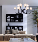 A modern multi-arm chandelier with a matte black metal frame and warm white glass shades hanging in a contemporary living room above a beige sofa.