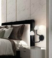 A modern black wall sconce with two curved arms and white dome-shaped shades, mounted on a light marble wall next to a bed with white and brown bedding.