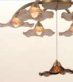 A close-up of a modern chandelier with multiple wavy, petal-like glass shades in a warm frosted beige finish and a lower pendant shade in a translucent amber-brown glass, all with brass-toned metal hardware against a plain off-white background.