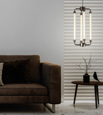 A modern multi-light pendant chandelier with four vertical frosted glass tubes and a dark metal frame, hanging in a contemporary living room next to a brown sofa and a striped window valance.