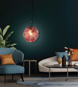 A modern pendant light with a faceted, translucent pink glass shade hangs in a stylishly decorated living room with a dark teal wall, blue armchair, and white sofa.