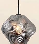 A modern pendant light with a ribbed, wavy metallic glass shade in a silver finish, illuminated from within, hanging against a plain beige background.