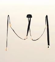 A modern black chandelier with three long hanging straps and circular cream-colored light diffusers, suspended against a plain off-white background.