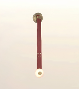 A minimalist wall-mounted pendant light with a circular gold-toned ceiling mount, a long vertical burgundy leather strap, and a glowing warm white spherical shade at the bottom, set against a soft cream background.