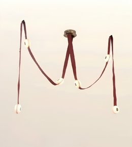 A minimalist modern pendant light fixture shaped like the letter 'M', featuring dark reddish-brown fabric straps and circular white ceramic discs, suspended against a solid pale beige background.