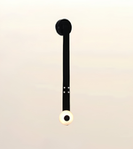 A minimalist black wall sconce with a circular black backplate and a spherical white globe shade featuring a black center, shown against a soft beige gradient background.