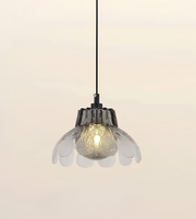 A modern pendant light with a black hanging cord and a flower-shaped clear glass shade, shown illuminated against a soft beige background.