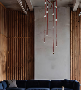 A modern living room interior featuring a dark navy blue sectional sofa at the bottom, flanked by vertical wooden slat walls on both sides and a textured light gray plaster wall in the center. A unique hanging chandelier with long red cords and white circular elements hangs from the ceiling.