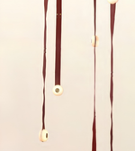 A minimalist wall hanging or mobile with multiple vertical dark red-brown ribbon-like strands and evenly spaced round cream-colored discs, shown against a plain light beige background.