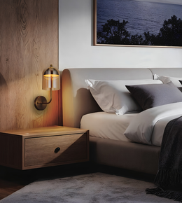 A modern bedroom scene featuring a bed with white and gray bedding, a matching wood nightstand, and a wall-mounted brass and glass bedside lamp. A wood-paneled accent wall and a framed landscape print are visible in the background.