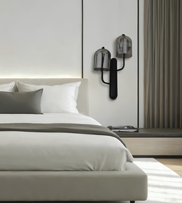 A modern minimalist bedroom featuring a low-profile platform bed with white bedding and gray accent pillows, a matching beige upholstered headboard and footboard, a built-in nightstand, and a pair of contemporary black wall sconces mounted on a white paneled wall.
