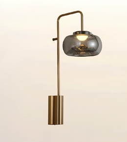 A modern wall sconce with a cylindrical brass base and an angled arm holding a smoked glass dome shade, shown against a plain light beige background.