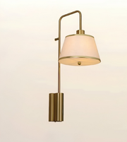 A modern wall-mounted sconce with a brushed brass/gold metal frame and a tapered off-white fabric shade, shown against a plain light background.