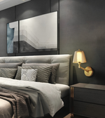 A modern bedroom scene featuring a bed with gray and white bedding, a lit gold wall sconce, and a dark gray wall with abstract framed wall art.