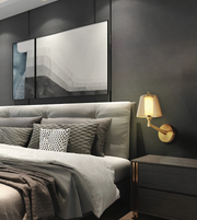 A modern bedroom scene featuring a bed with gray and white bedding, a lit gold wall sconce, and a dark gray wall with abstract framed wall art.