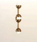 A modern vertical wall sconce with a brushed brass/gold finish, featuring three stacked semi-spherical metallic sections with integrated white diffusers, shown against a soft beige gradient background.