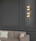 A modern wall sconce with three stacked cream-colored glass shades and polished gold-toned metal hardware, mounted on a dark gray paneled wall above a beige sofa with white pillows.