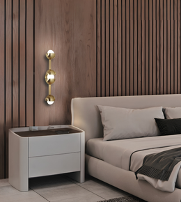 A modern bedroom scene featuring a wall-mounted three-bulb gold sconce on a dark wood slatted wall, next to a low white nightstand and a beige upholstered bed.